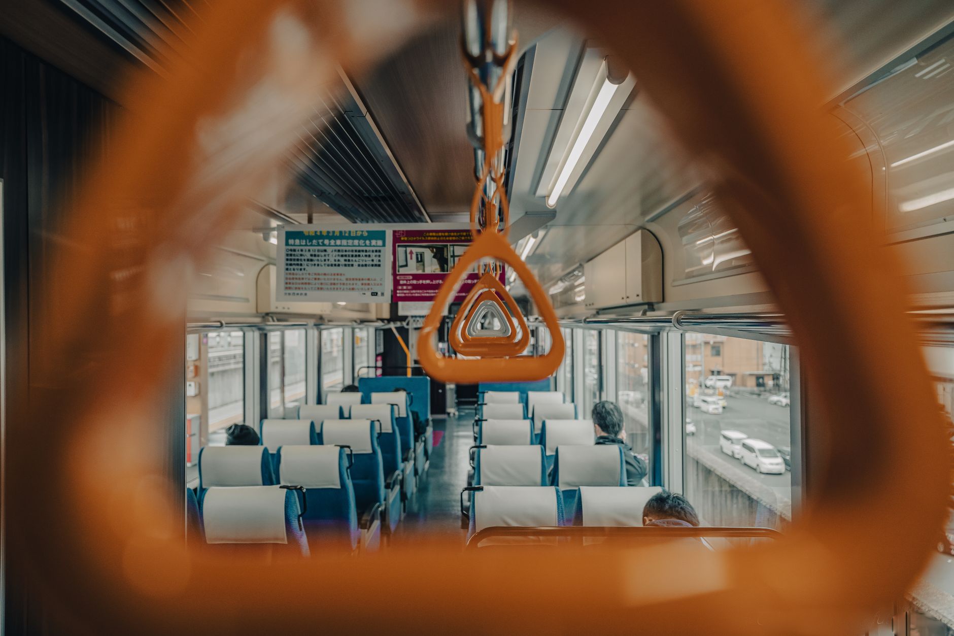 電車の車内。オレンジ色のつり革の向こうに、座席と窓の景色が続いている。