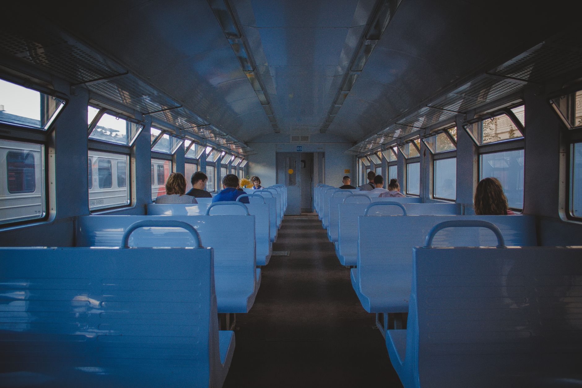 青い座席が並ぶ電車の車内で、数人の乗客が席に着いているところ。