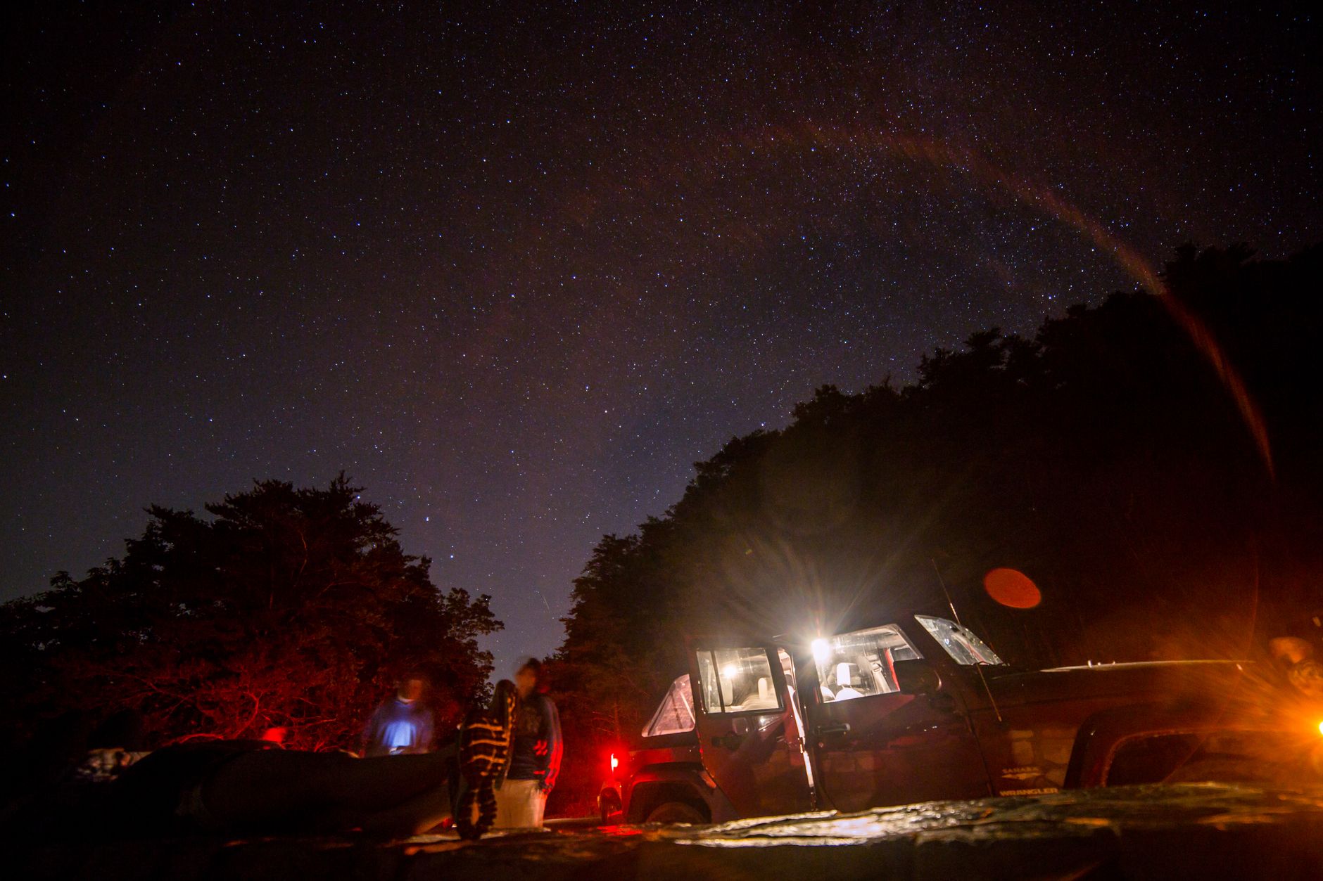 星空の下のキャンプ地で、ドアを開けて室内灯を点けた車と、その周りに立つ人。