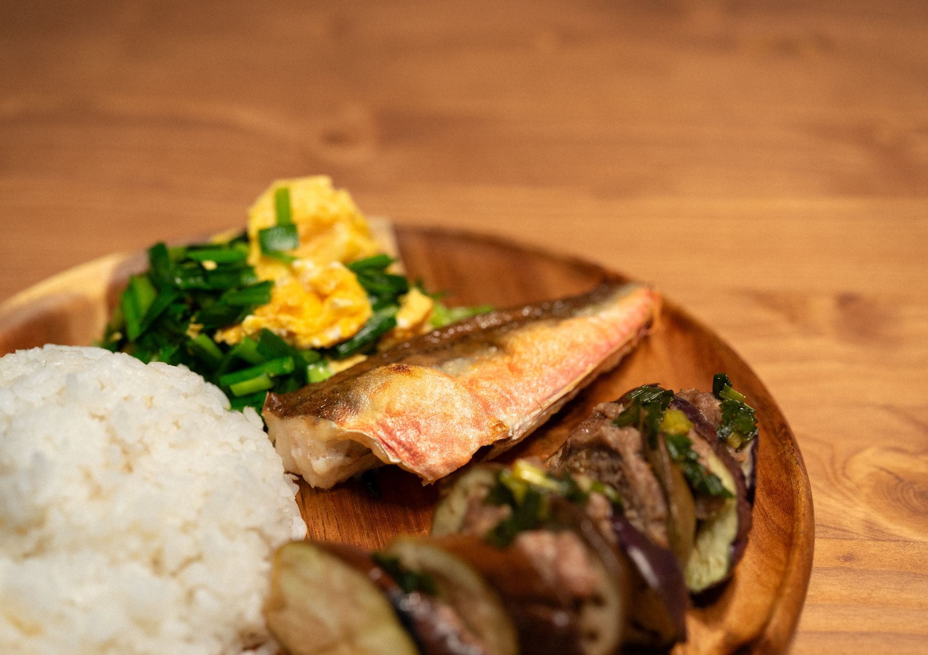 木の皿に白いごはんと焼いた魚、青菜と卵のおかずを盛りつけた家庭の献立。