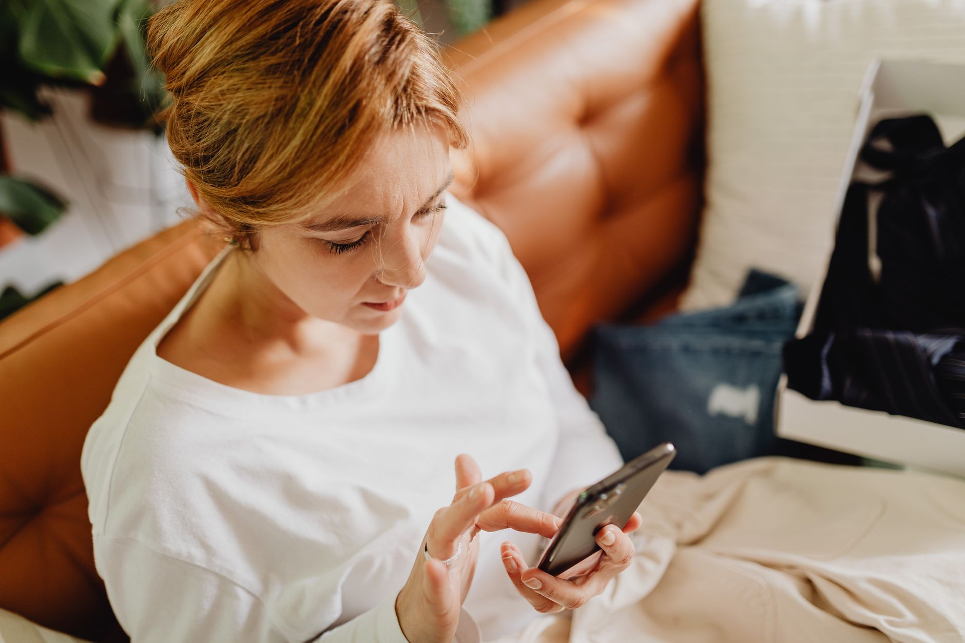 部屋のソファに座り、手元のスマートフォンを見て考え込んでいる女性。