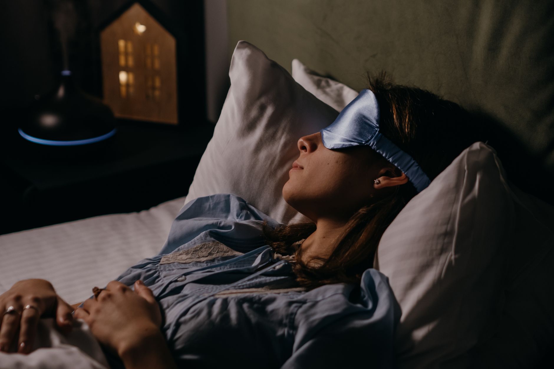アイマスクを着けて眠る人。間接照明だけがともる、夜のベッドまわり。