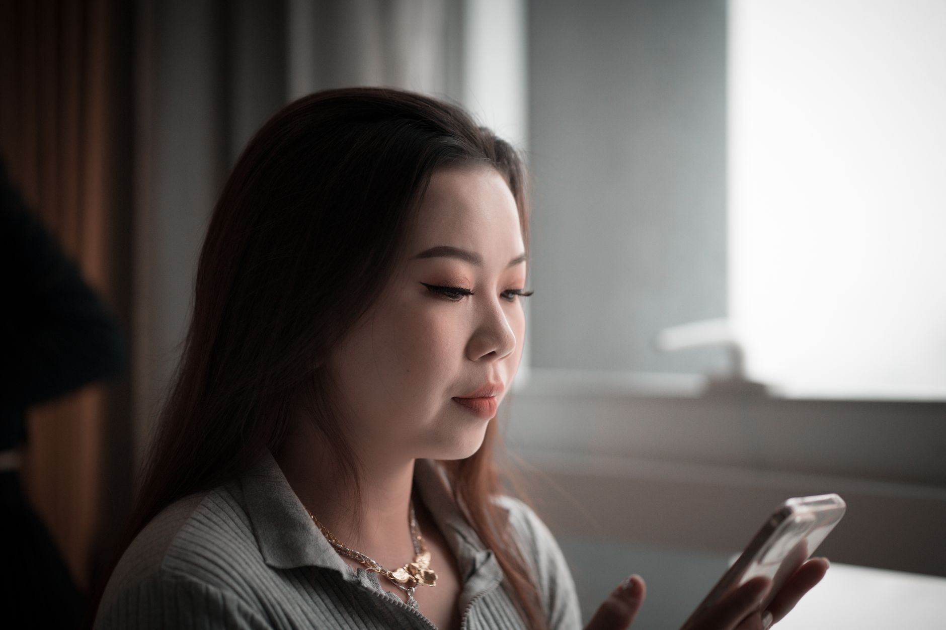 窓辺の明るい室内で、手に持ったスマートフォンの画面に目を落とす女性。