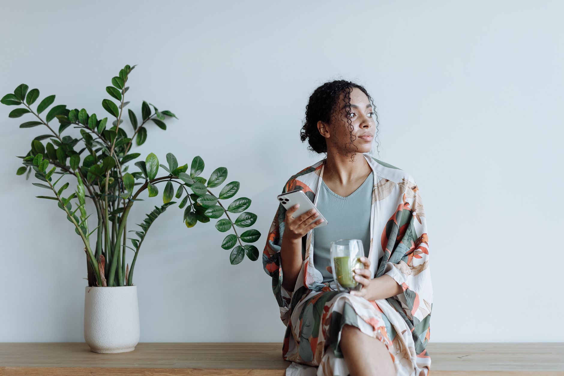 観葉植物のそばに腰かけ、飲み物とスマートフォンを手に考えごとをする人。