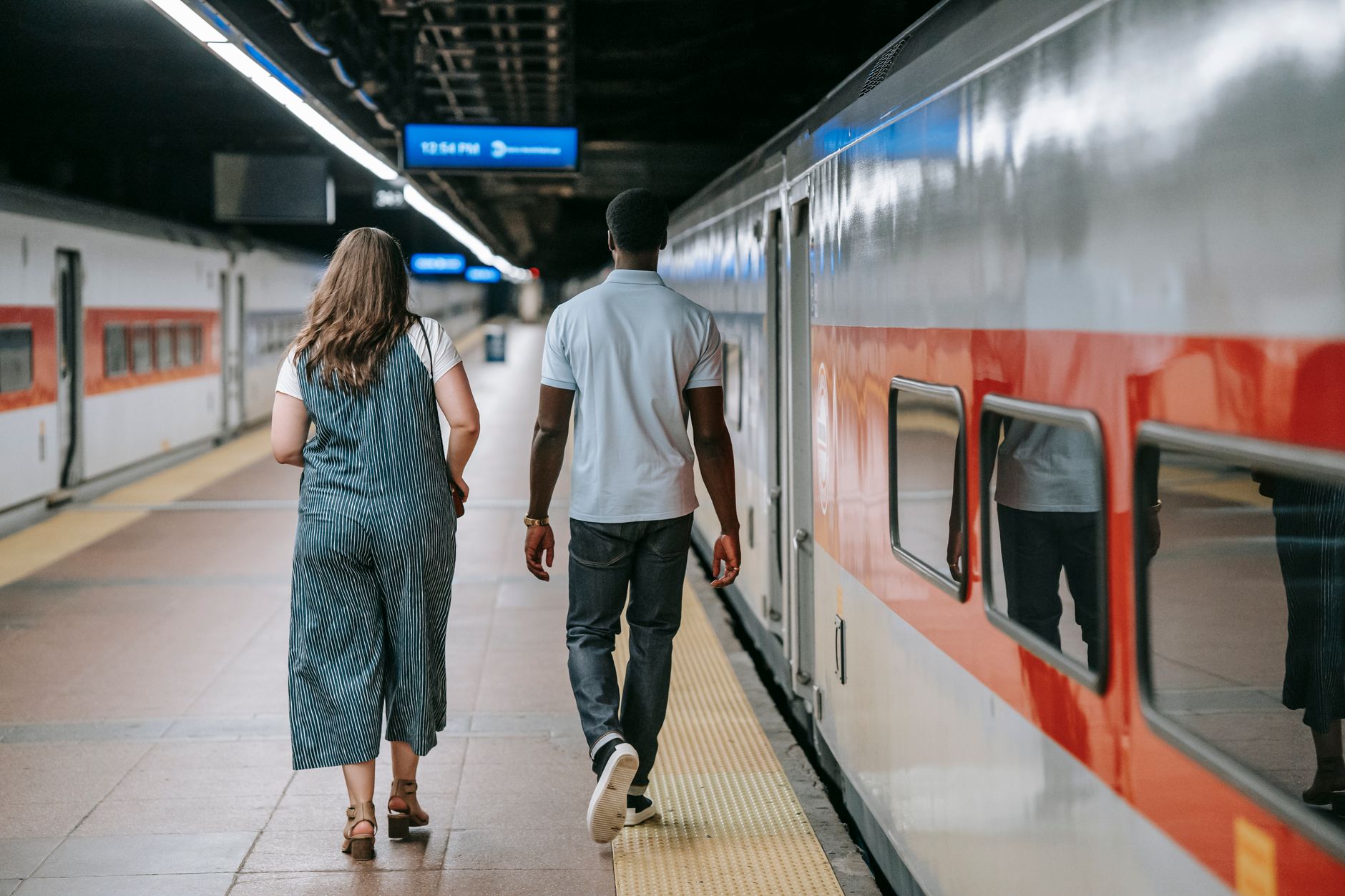夏の装いの男女が、停車中の列車の脇のホームを並んで歩いている後ろ姿。