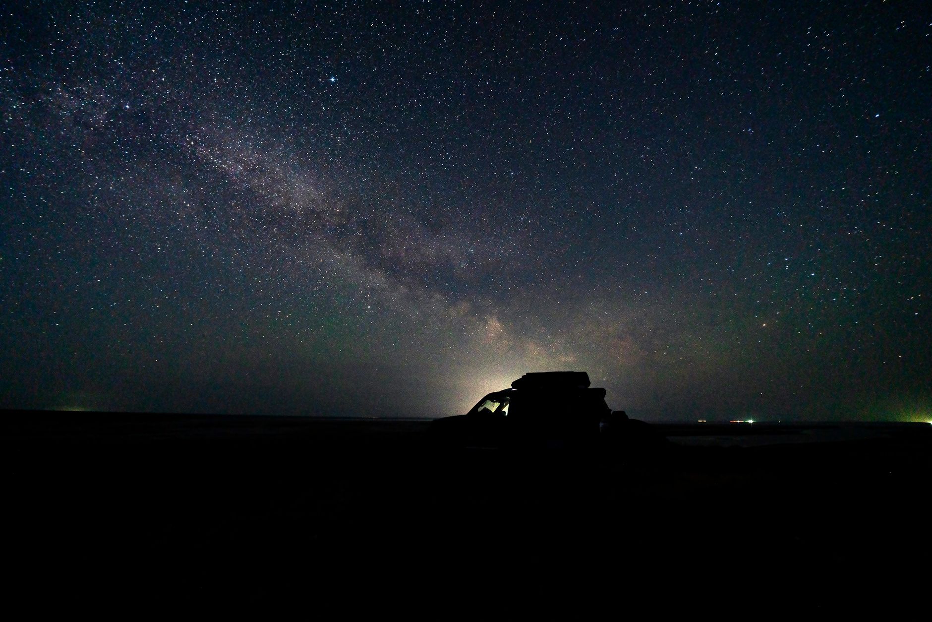 星空の下、暗い野原に停めた車の輪郭だけが浮かび上がっている夜の風景。